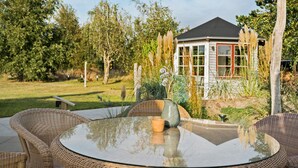 A glass-top table with wicker chairs in a garden setting, a small potted plant, and a house with a dark roof and white walls in the background.