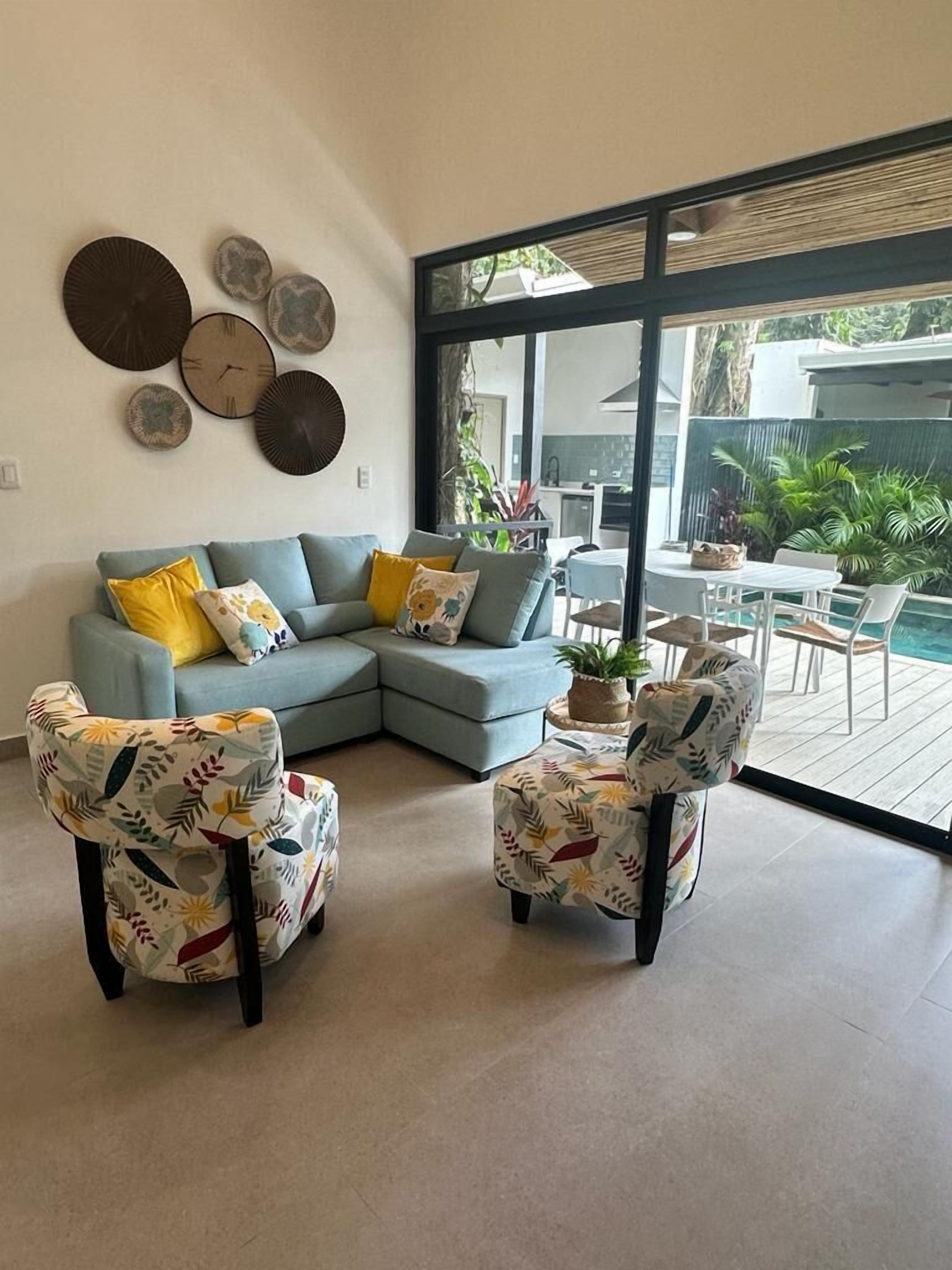 A modern living room with a light blue sectional sofa, two patterned armchairs, and a coffee table. There are decorative plates on the wall and a view of a patio with outdoor furniture.