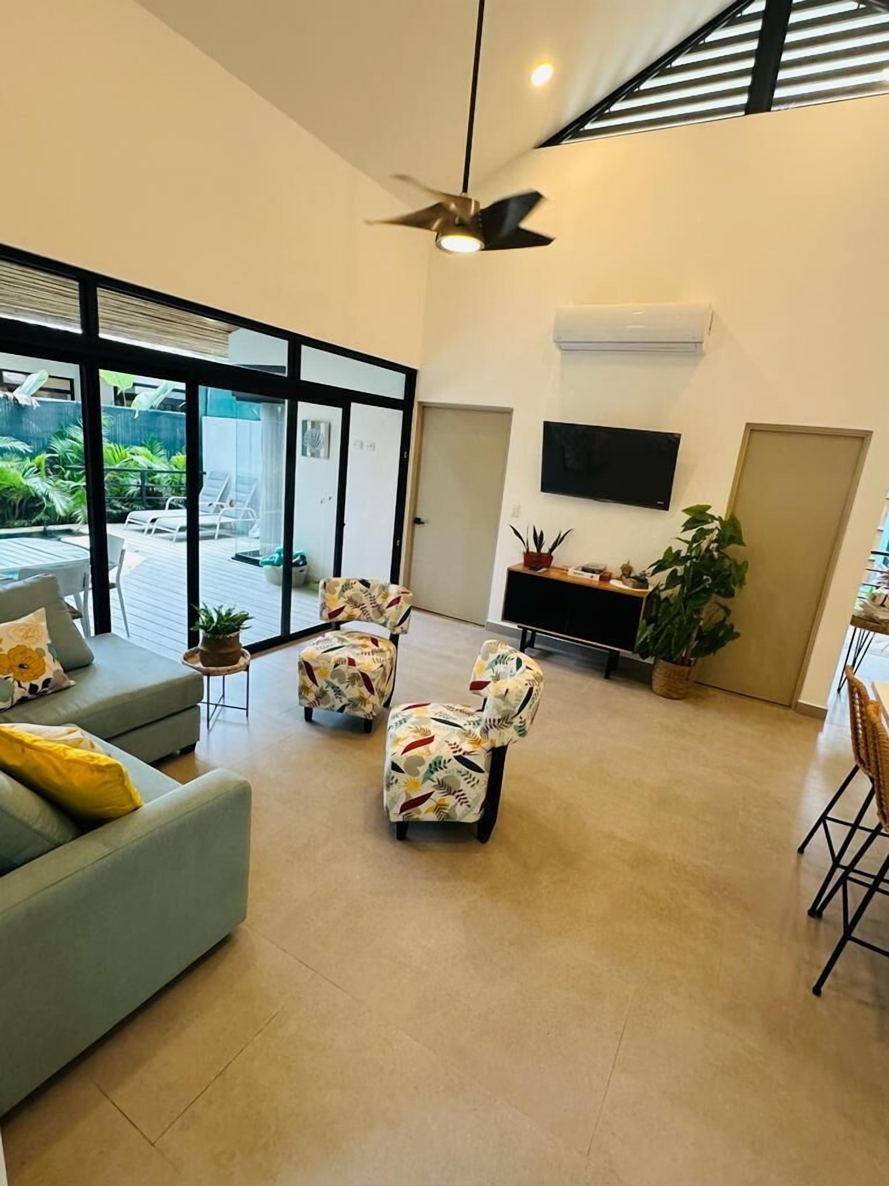 A spacious living area with a grey sofa, floral-patterned chairs, a ceiling fan, a flat-screen TV, and a view of the outdoors through large windows.