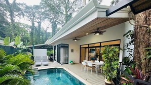 A modern outdoor pool area with a wooden deck, a dining table with chairs, and lush greenery. - Casa Coral Azul (Cahuita)