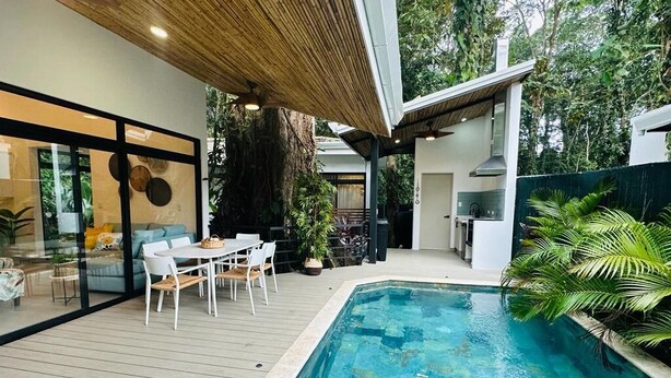 A modern outdoor pool area with a wooden deck, a dining table with chairs, and a kitchen area. - Casa Coral Azul (Cahuita)