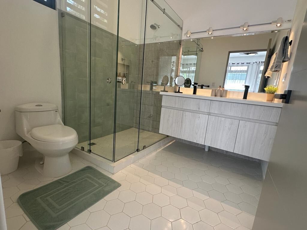 A modern bathroom with a glass-enclosed shower, a white toilet, a vanity with a mirror, and a green rug.