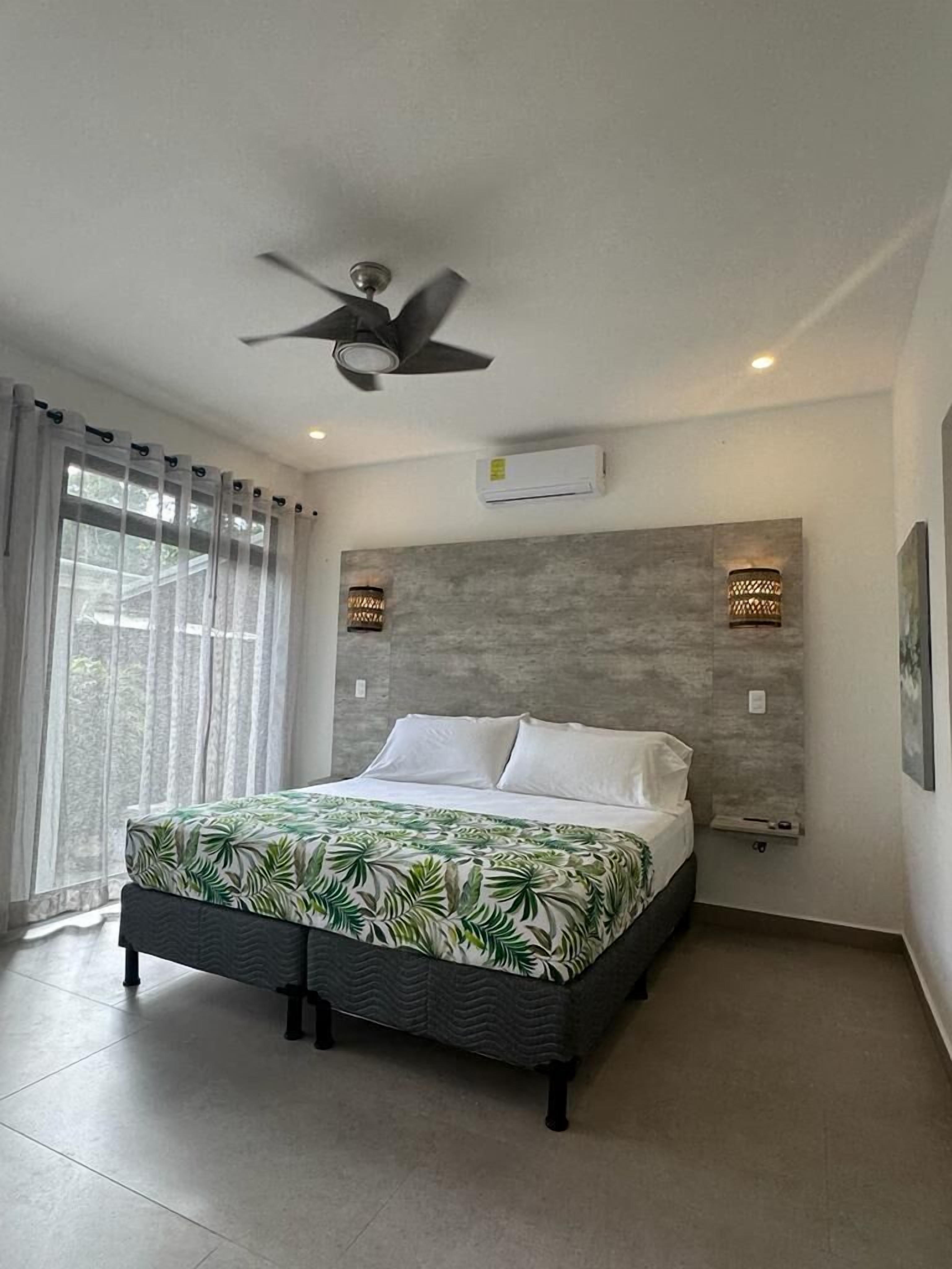 Une chambre moderne équipée d’un lit, d’un ventilateur de plafond, d’un appareil de climatisation et de deux lampes fixées au mur.