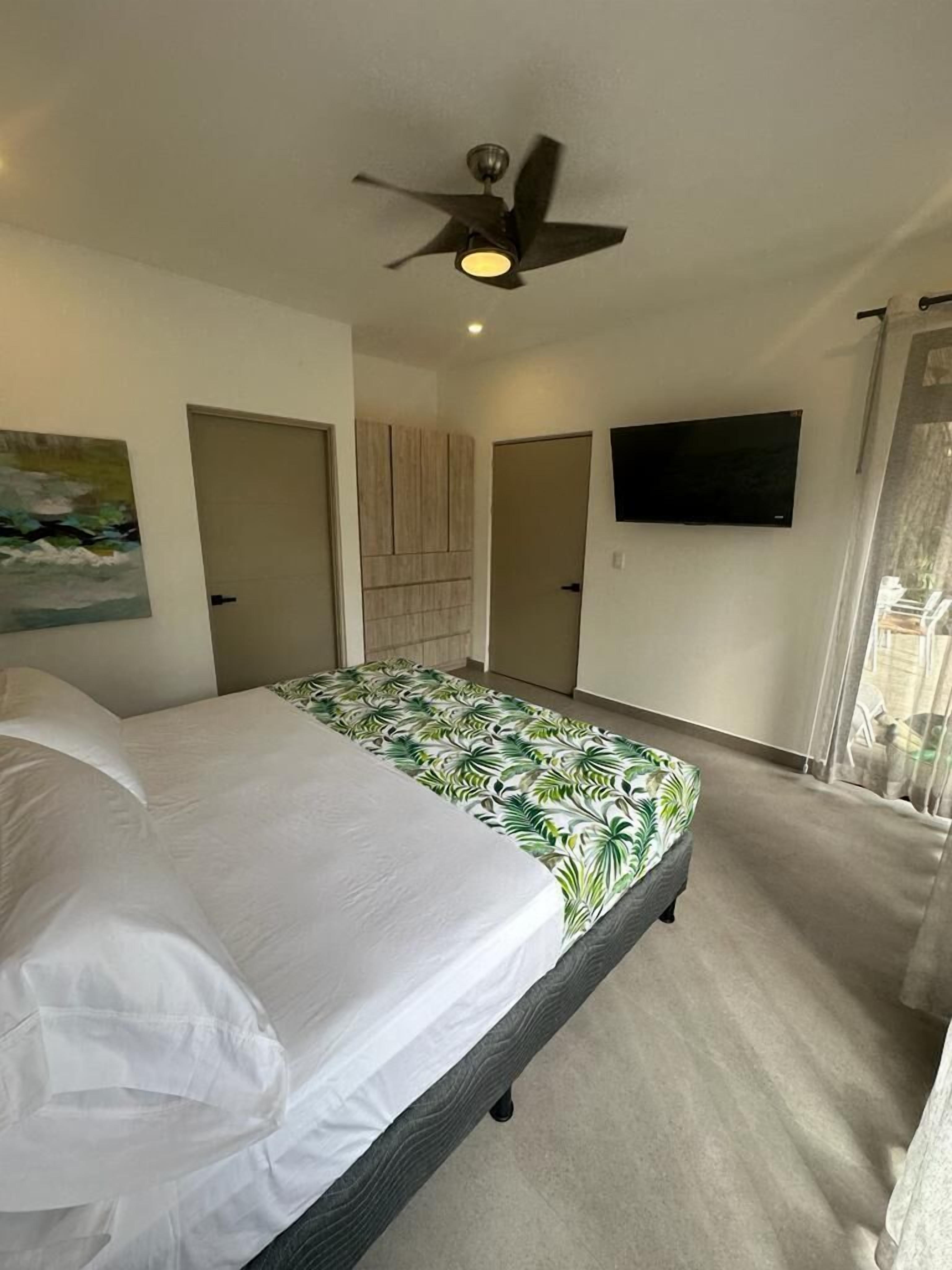Un dormitorio con cama, ventilador de techo, televisor y un cuadro en la pared.