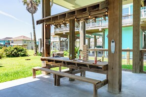 A wooden outdoor seating area with a picnic table and benches, surrounded by a wooden deck and a view of a residential area with houses and a palm tree. - Steps to Beach, Hot Tub, Gulf & Beach Views, Tiki Bar, Grill, Dogs, Games (Bolivar Peninsula)