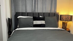 A bedroom with a large bed, grey curtains, a nightstand with a lamp, and a wooden side table.