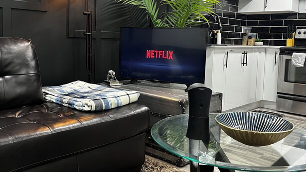 A modern living room with a leather sofa, a glass coffee table, a television displaying 'NETFLIX', a floor lamp, and a kitchen area in the background.