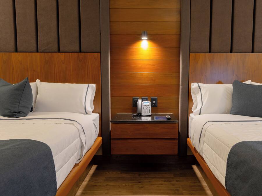 A hotel room with two beds, a nightstand with a phone and a coffee maker, and a wooden panel wall with a light fixture.