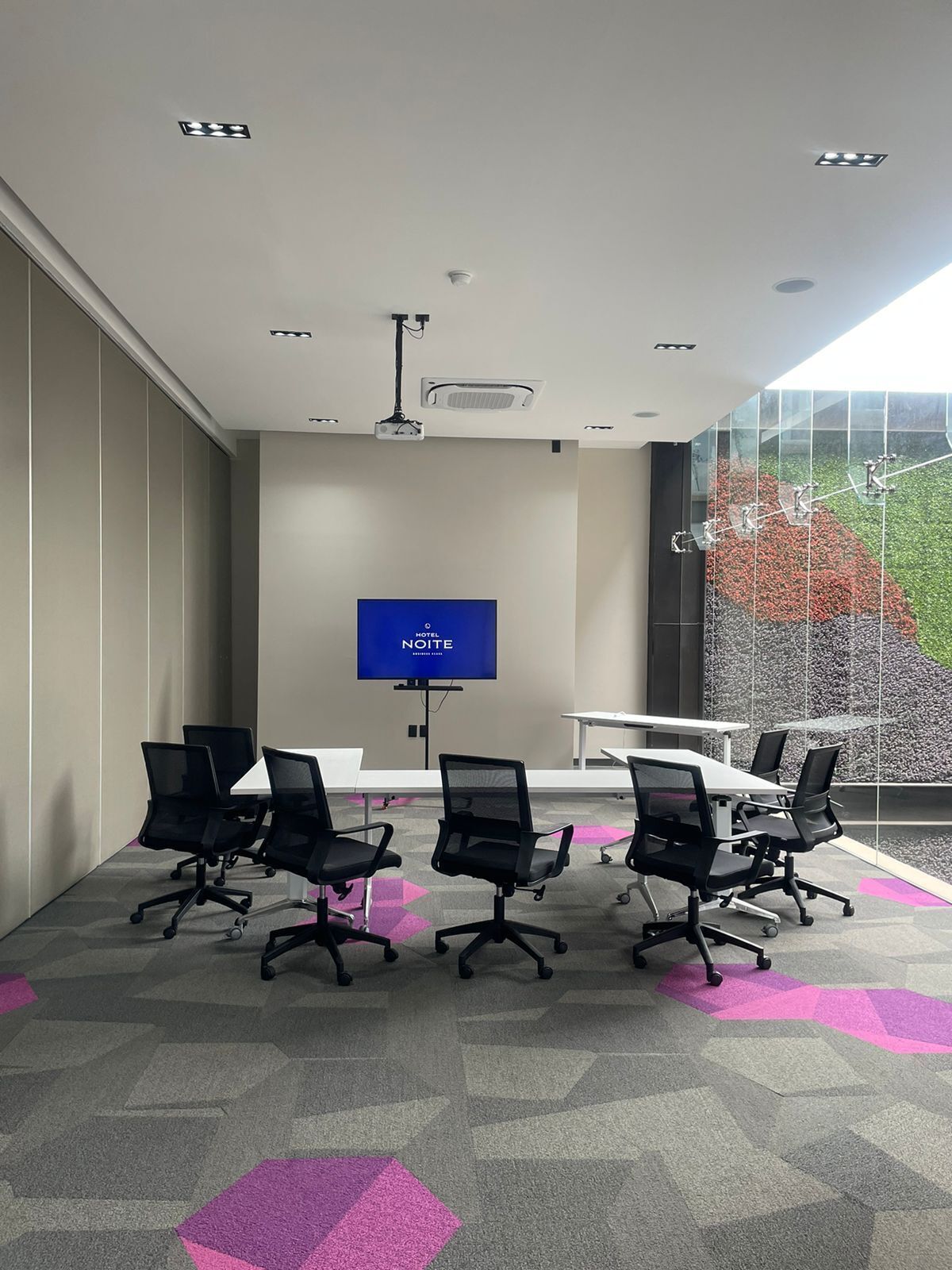 A modern conference room with a large flat-screen TV, black office chairs, a white table, and a patterned carpet.