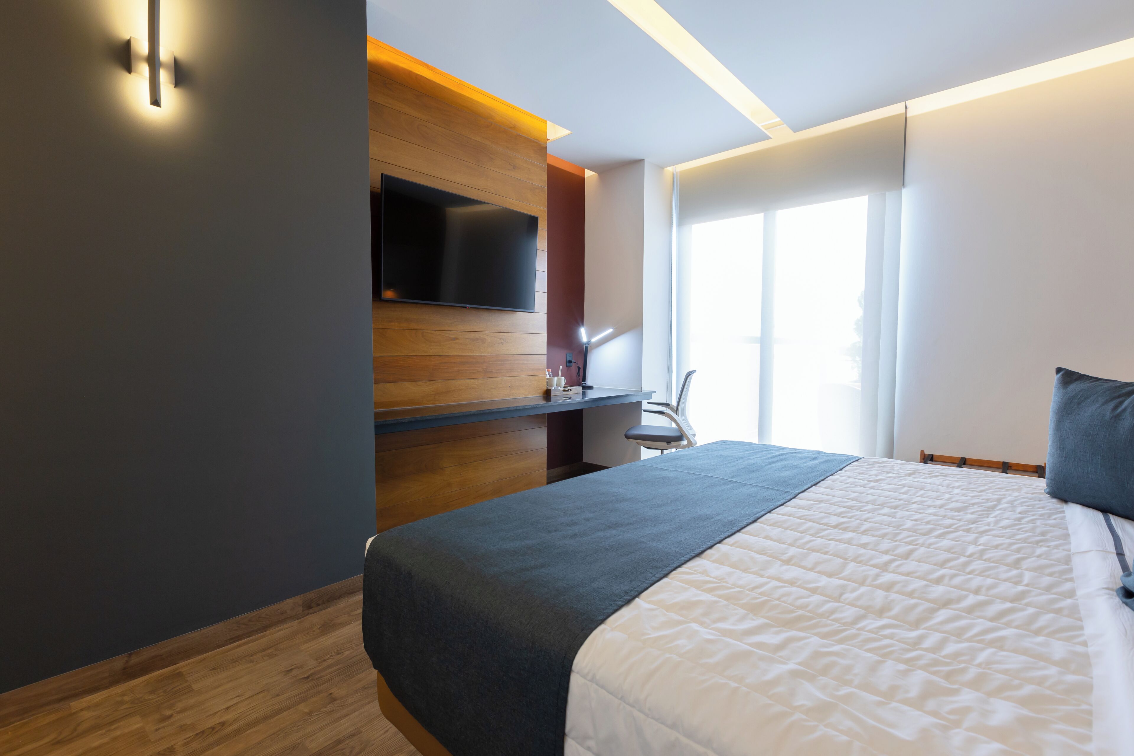 A modern hotel room with a large bed, a flat-screen TV mounted on a wooden panel wall, a desk with a chair, and a large window providing natural light.