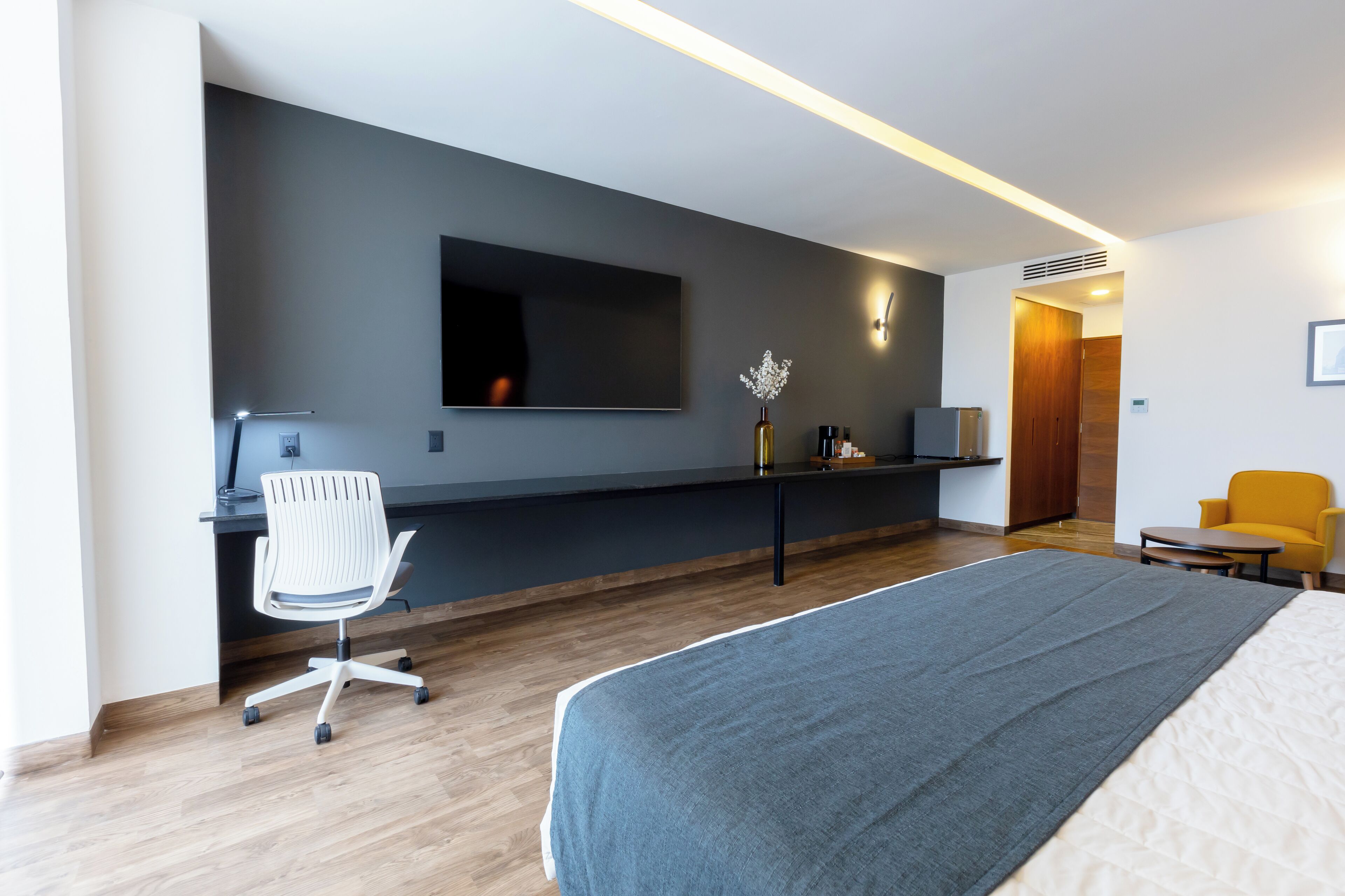 Habitación de hotel moderna con una cama grande, un escritorio con silla, un televisor de pantalla plana y un pequeño rincón de descanso con una silla amarilla.