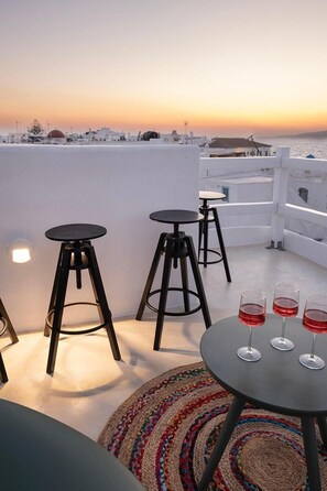 Una terraza en la azotea con tres taburetes negros, una mesa redonda con tres copas de vino y una alfombra tejida de colores.