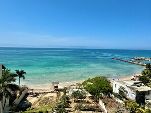 Vista costera con playa, agua azul cristalina y un rompeolas rocoso.