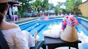 Une personne se détend au bord d’une piscine, assise sur une chaise avec une serviette et un sac en tissu à ses côtés.