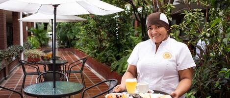 A waiter holding a tray with food and drinks in an outdoor patio area with tables and chairs.