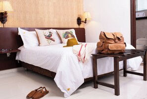 Una cama bien hecha con una colcha blanca, un cabecero marrón y almohadas decorativas. Un bolso de cuero marrón está sobre una mesita de noche de madera junto a la cama.