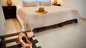 Un lit bien fait, recouvert d’une couvre-lit beige, et sur lequel est posé un chapeau jaune. Il y a deux tables de chevet avec des lampes, et une paire de chaussures est posée sur un banc devant le lit.