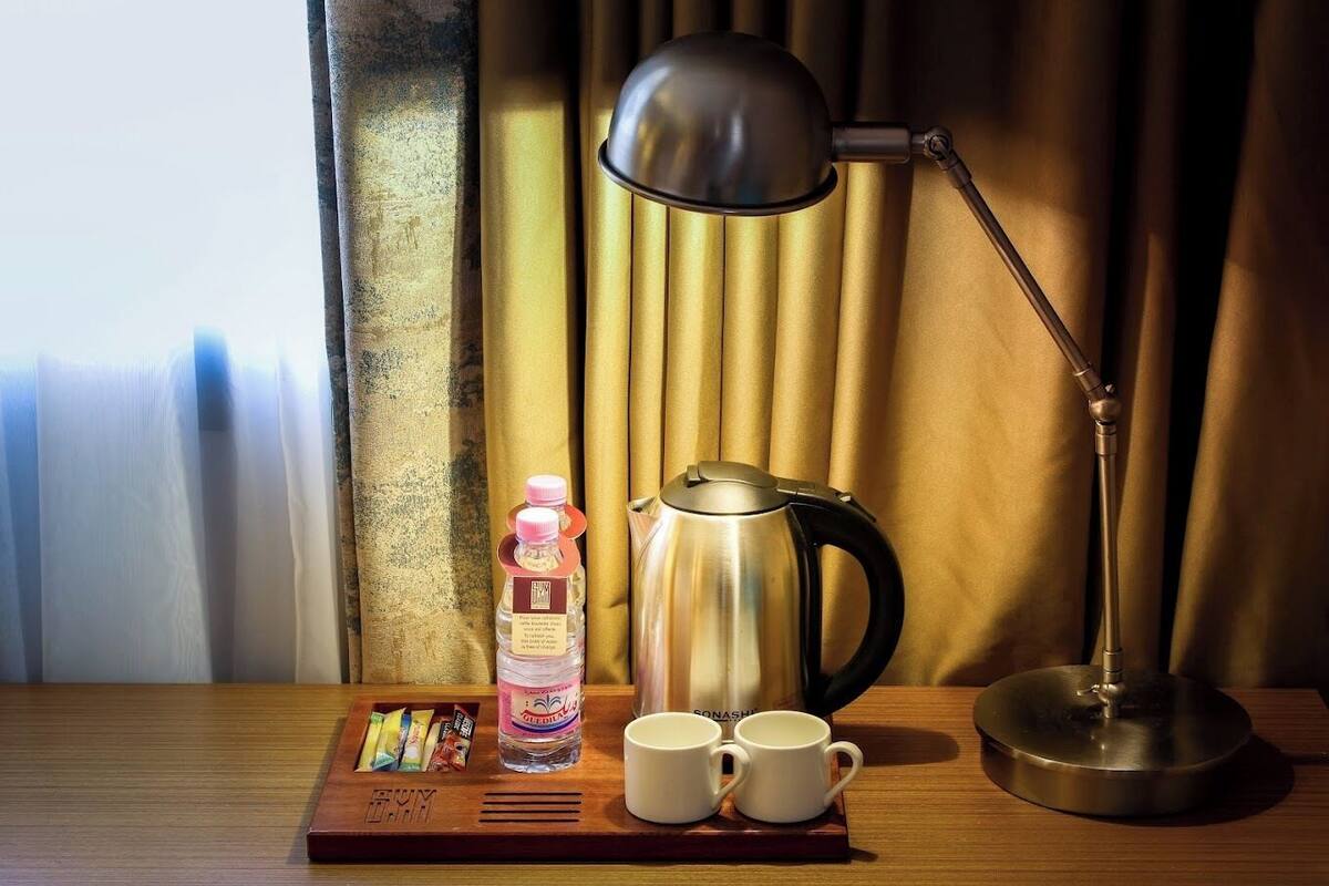 A wooden tray with a water bottle, two white mugs, a stainless steel kettle, and a black desk lamp on a wooden table.
