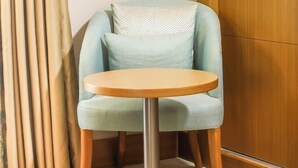 A corner room with a light blue armchair, a round wooden table, beige curtains, and a wooden wardrobe. - Sym Hotel - Chéraga (Algiers)