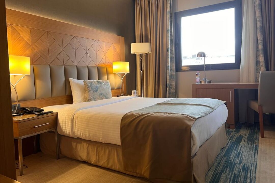 A hotel room with a large bed, two bedside lamps, a desk with a chair, and a window with curtains.