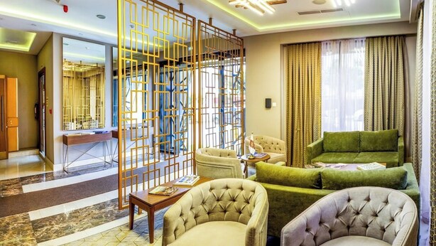 A hotel room with a seating area featuring a green sofa, two armchairs, and a coffee table. There is a decorative screen with geometric patterns and a large mirror on the wall. - Sym Hotel - Chéraga (Algiers)