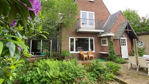 Une maison à deux étages, construite en briques, avec un balcon, un jardin agrémenté de fougères et des meubles d’extérieur.