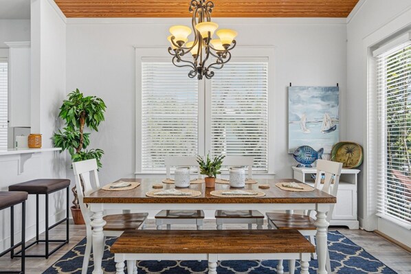 A dining area with a wooden table, white chairs, a chandelier, and a view through the window. - SEA-REST!! Relaxed 4br 4ba home, POOL and close to the BEACH! (Alys Beach)