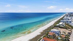 Une vue aérienne d’une zone côtière, avec une plage, des eaux d’un bleu limpide et une rangée de bâtiments présentant un style architectural uniforme.