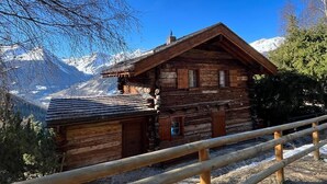 Una cabaña de madera con techo inclinado, rodeada por una valla de madera, con montañas nevadas y un cielo azul despejado al fondo.