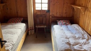 Una habitación de madera con dos camas, una silla, un ventanal y una puerta.