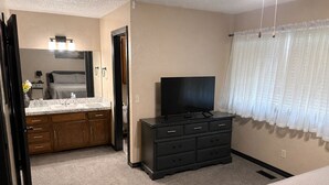 Une chambre à coucher comprenant un lit, une commode, un téléviseur, une salle de bain avec un lavabo et un miroir, et une fenêtre avec des rideaux.