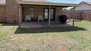 Une maison en briques avec un patio couvert, deux chaises, une table et un barbecue.