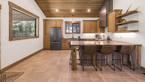 Une cuisine moderne dotée d’un grand îlot central, de meubles en bois et offrant une vue sur la forêt par une fenêtre.