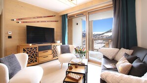 A modern living room with a large TV, wooden furniture, and a view of a snowy landscape. - Ski aux pieds & piscine | T2 + cabine (Alpe d'Huez)