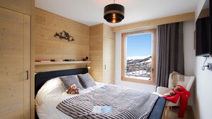 A bedroom with a bed, a window with a view, wooden walls, and a television. - Ski aux pieds & piscine | T2 + cabine (Alpe d'Huez)