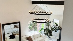 A spacious living room with a white sofa, two armchairs, a glass coffee table, and a large mirror. A modern chandelier hangs from the ceiling, and a staircase with a black railing is visible. - w/pool 5 min to the Strip (Las Vegas)