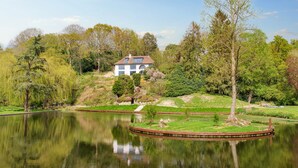 A serene lakeside setting with a house on a hill, surrounded by lush greenery and trees. - 5 Bed Lake House - Sleeps 10 - Nature Reserve (Devizes)