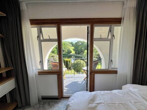 A bedroom with a large sliding glass door leading to a balcony with a view of greenery and a pond.