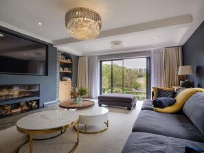 A modern living room with a large TV, a chandelier, a sofa, and a view of greenery outside.