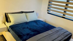 Ein ordentlich bezogenes Bett mit einer blau-grauen Bettdecke, ein weißer Nachttisch mit Lampe und ein Fenster mit horizontalen Jalousien.