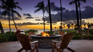 Un foyer extérieur entouré de quatre chaises en bois, le tout niché au milieu d’une végétation luxuriante et de palmiers, avec vue sur une plage au coucher du soleil.