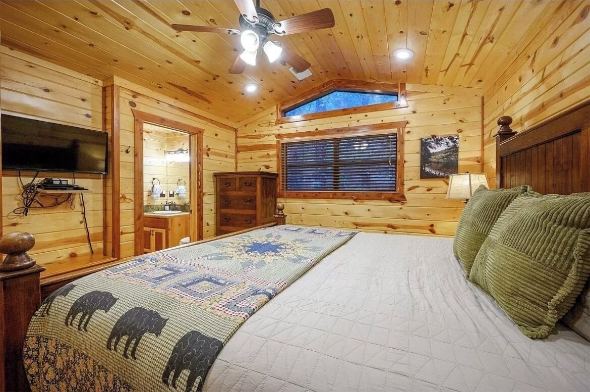 A log cabin bedroom with a bed, a ceiling fan, a window with blinds, a TV mounted on the wall, and a wooden cabinet.