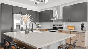 A modern kitchen with dark cabinets, a large island with bar stools, stainless steel appliances, and a decorative vase. - Beautifully Designed, Fully Stocked New Condo with Custom Features - Plus A Gym! (Columbia Falls)