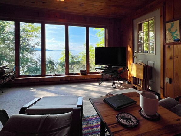 A wooden cabin interior with a large window overlooking a lake, a television on a stand, and a dining table with chairs.