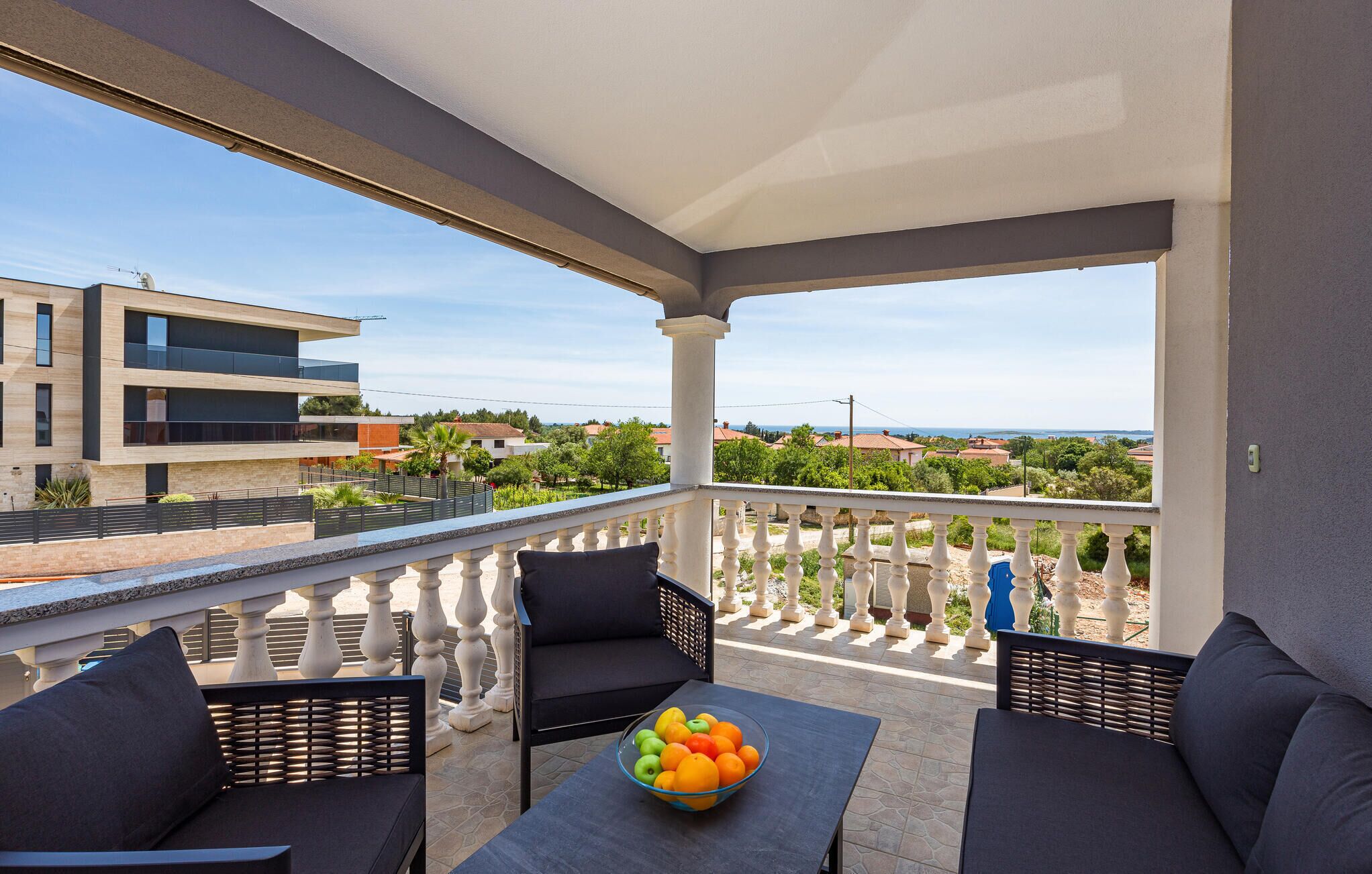 A balcony with a view of a modern building, a bowl of fruit on a table, and two chairs.