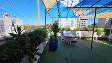 A rooftop terrace with a table and chairs, surrounded by potted plants and a blue and yellow canopy.
