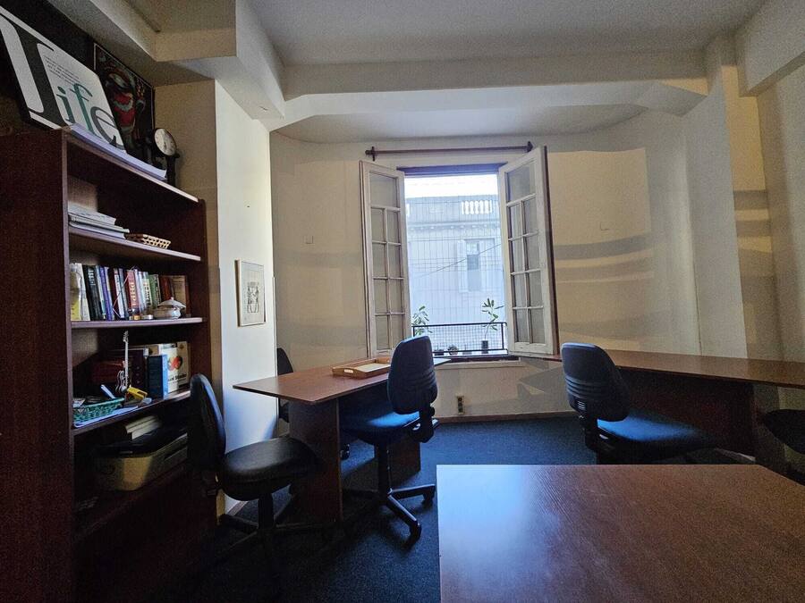 A room with a desk, two chairs, a bookshelf, and a window with a view of buildings.