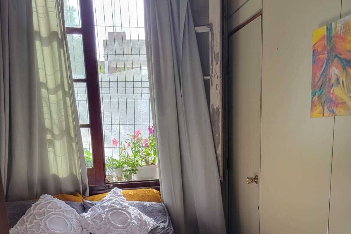 Quarto com cama, cortinas, janela com plantas, guarda-roupa e um tapete colorido.