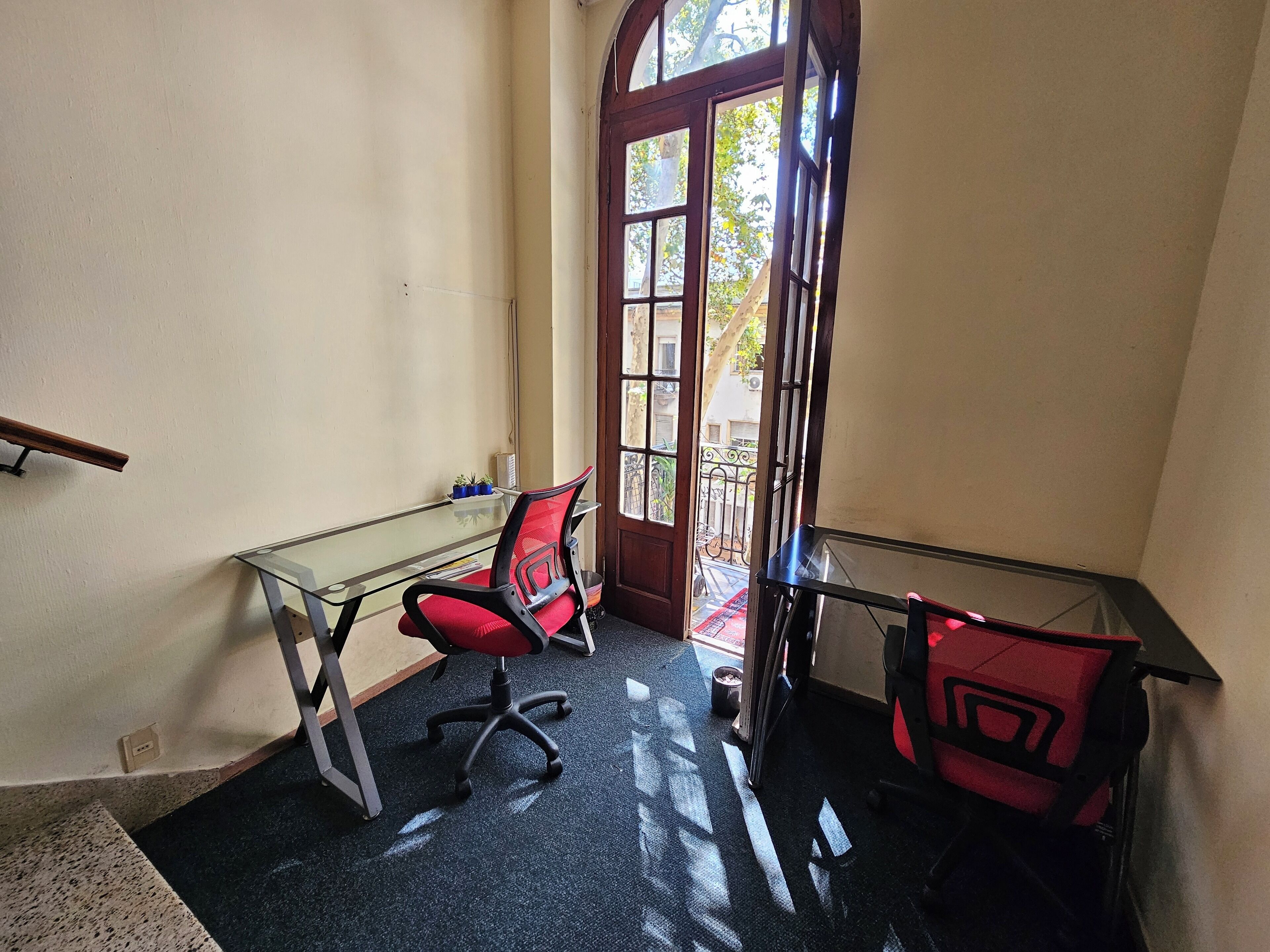 Un pequeño espacio de oficina con un escritorio de vidrio, dos sillas de oficina rojas, una silla de oficina negra y un banco de madera fuera de la puerta.