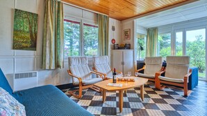 Een knus vertrek met een houten plafond, een geruite vloer, een bank, twee fauteuils, een tafeltje met een fles en glazen, en grote ramen met gordijnen.